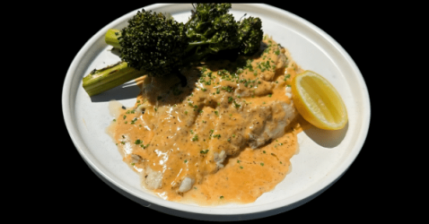 A white plate with fish fillets covered in creamy orange sauce, garnished with chopped herbs, served with steamed broccolini and a lemon wedge.