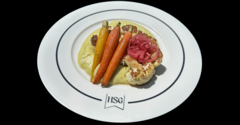 A white plate with the initials HSG holds colorful roasted carrots, a cauliflower floret, pink pickled onions, and a creamy yellow purée, all arranged neatly against a black background.