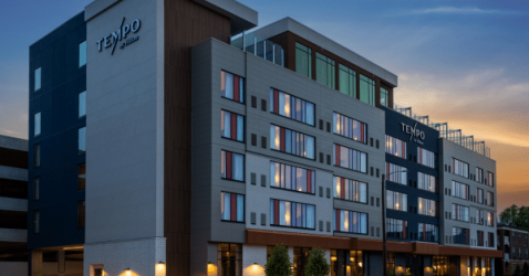 A modern, multi-story Tempo by Hilton hotel exterior at dusk, with large windows lit from within and the hotel name displayed prominently on the building.