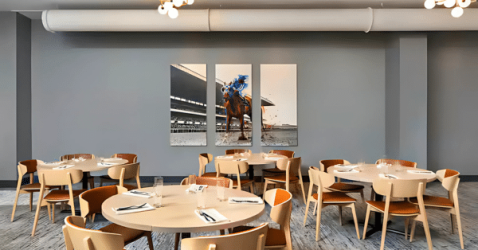 A modern dining area with round tables and wooden chairs, each table set with napkins and glasses. Three-panel artwork of a racehorse decorates the gray wall. Ceiling lights and exposed ductwork are visible above.