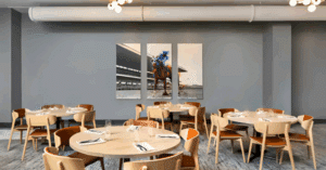 A modern dining area with round tables and light wooden chairs, set for guests. Three-panel horse race artwork adorns a gray wall. Ideal for an Interactive dinner theater in NuLu or The Dinner Detective Louisville at Tempo by Hilton NuLu.