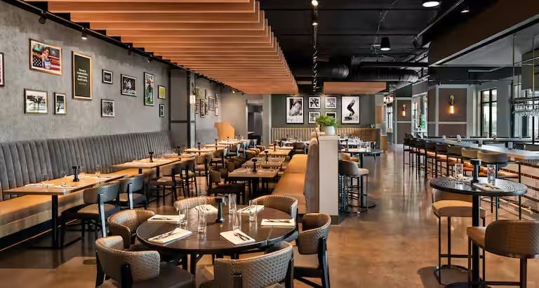 Modern restaurant interior with wooden tables, cushioned chairs, and a long booth along a wall decorated with framed art. The space has a warm, inviting atmosphere and an open layout with a bar area on the right.