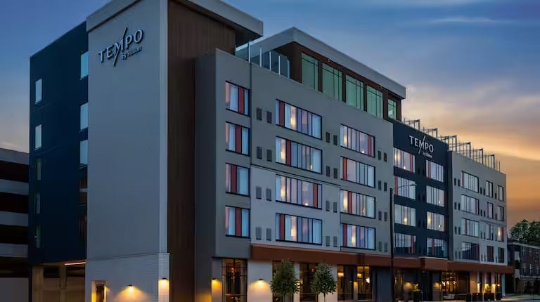 Modern multi-story hotel building with Tempo by Hilton signs, large windows on each floor, and soft exterior lighting at dusk, with a clear sky in the background.