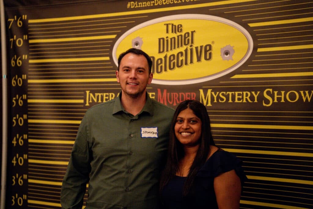 A smiling man and woman stand in front of a “The Dinner Detective: Interactive Murder Mystery Show” backdrop, perfect for private events in Louisville. The man wears a green shirt with a nametag, and the woman wears a dark dress.