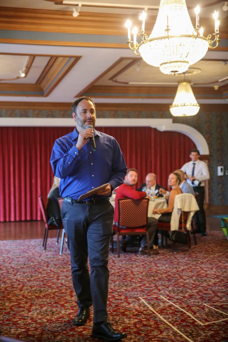 Host speaking during a Dinner Detective murder mystery show in a Louisville ballroom, with guests seated at round tables under chandeliers