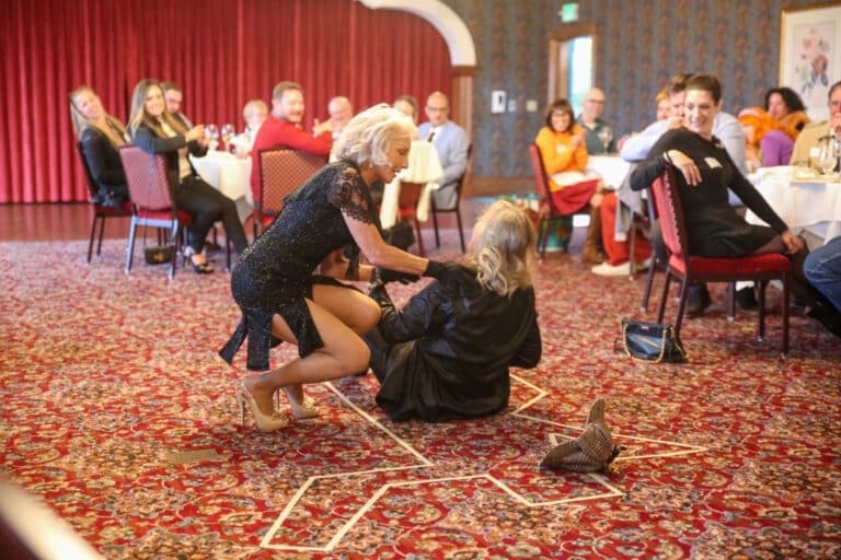 Guests laugh as two performers stage a dramatic clue reveal on the banquet room floor during a Louisville immersive dinner theater show in NuLu