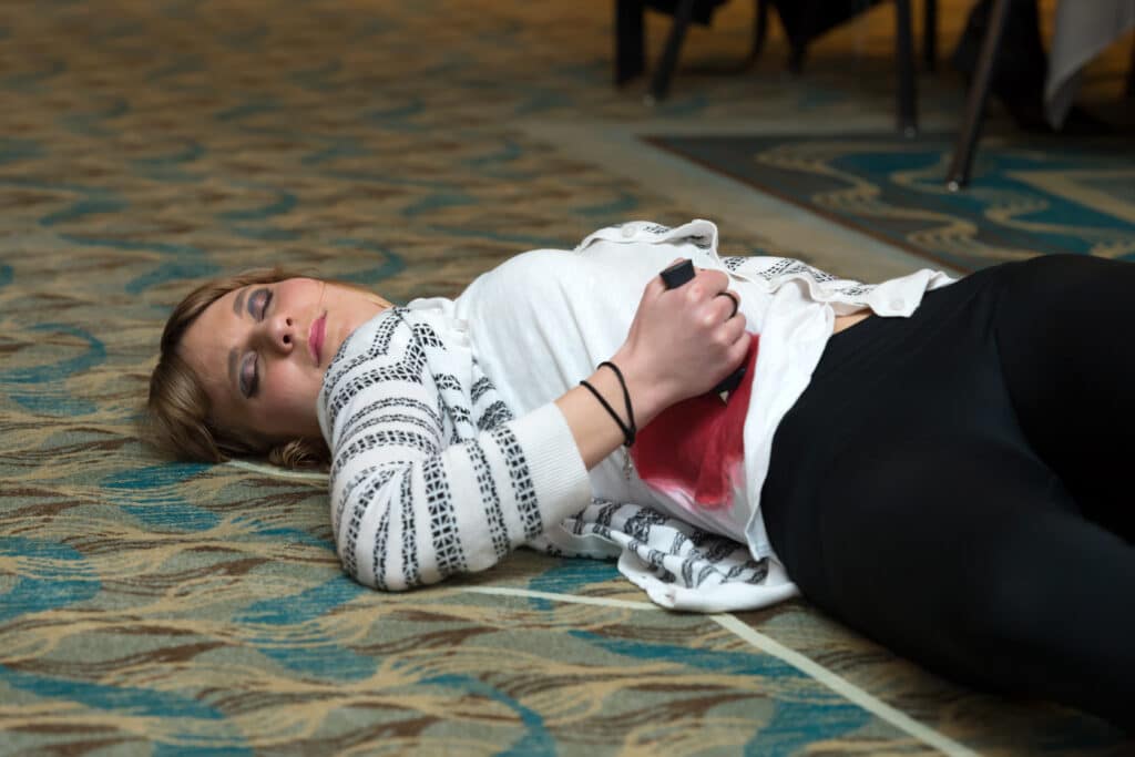 Actor playing the victim in a Dinner Detective murder mystery show, lying on the ballroom floor with comedic “crime scene” props