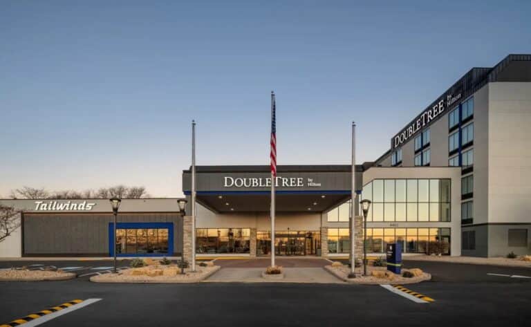 Front view of a DoubleTree by Hilton hotel at sunset, featuring a central entrance, American flag, large glass windows, and the Tailwinds restaurant on the left side. The sky is clear and the driveway is empty.