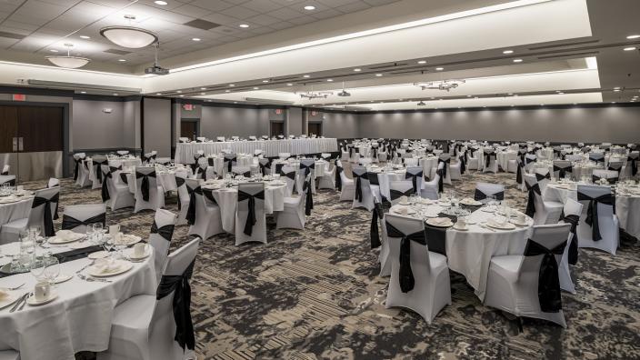 A large banquet hall set up for an event, featuring many round tables covered with white tablecloths and black chair sashes. Each table is set with plates, glasses, and napkins. The room has neutral walls and ceiling lights.