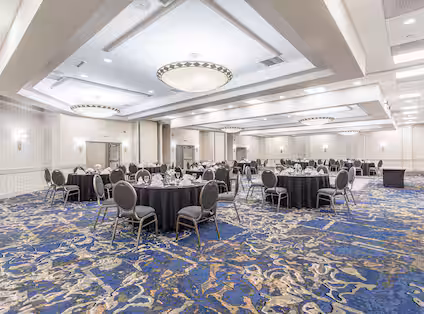 A spacious, elegant banquet hall with round tables covered in black tablecloths and surrounded by gray chairs, set on a blue patterned carpet, under large, modern chandeliers.