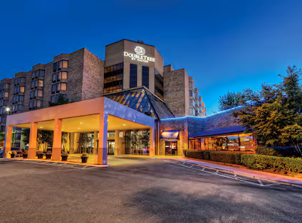 Exterior view of a DoubleTree hotel at dusk with bright lighting, a covered entrance, large windows, and surrounding greenery under a deep blue sky.