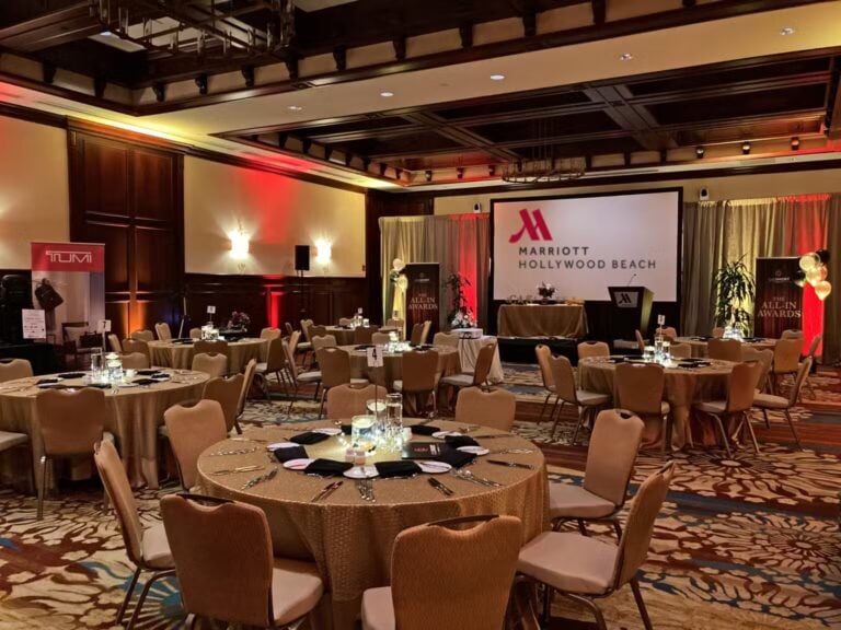 A banquet hall with round tables set for an event, featuring elegant table settings, candle centerpieces, and a large screen displaying Marriott Hollywood Beach. Warm lighting creates a welcoming atmosphere.