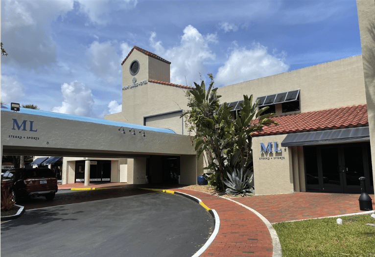 A beige building with red roof tiles and a clock tower displays ML and Stages & Sports signs. There are tropical plants, a curved driveway, and a brick walkway under a partly cloudy sky.