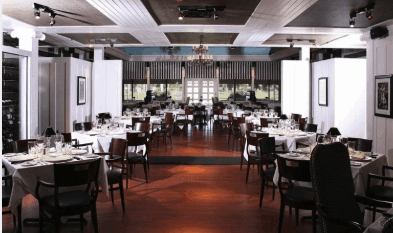 Elegant restaurant dining room with wooden floors, white tablecloths, and neatly set tables and chairs. Large windows at the back let in natural light, and a chandelier hangs from the ceiling. The room looks spacious and inviting.