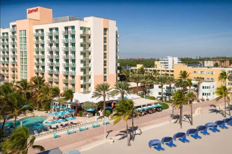A beachfront Marriott hotel with balconies overlooks a pool, palm trees, and rows of blue lounge chairs on a sandy shore under a clear blue sky.