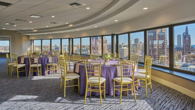 A banquet room with round tables covered in purple tablecloths and surrounded by gold chairs, set against large windows offering a city skyline view on a sunny day.