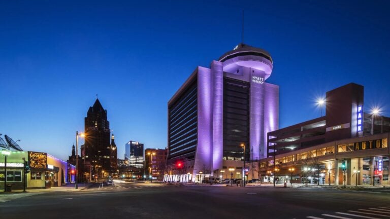 Street view of a city at dusk featuring a tall, modern building lit with purple lights, a parking structure to its right, and other illuminated buildings in the background under a deep blue sky.