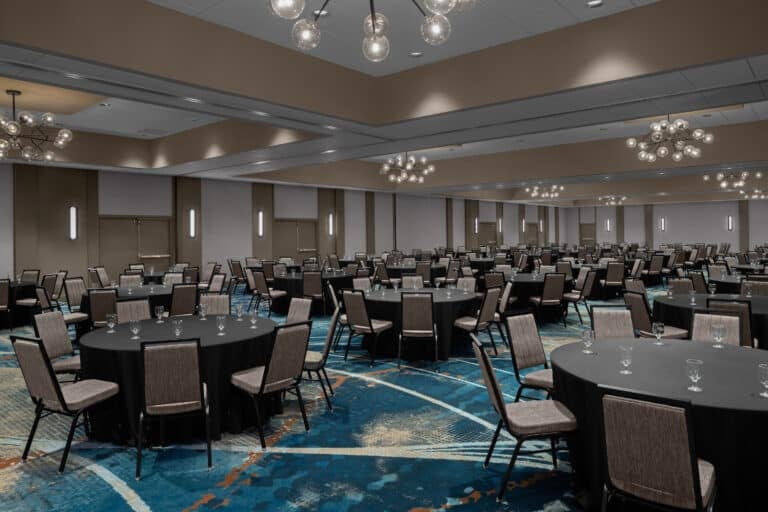 A large, modern banquet hall set up for an event with round tables covered in black tablecloths, surrounded by chairs, each table decorated with glassware; contemporary chandeliers hang from the ceiling.