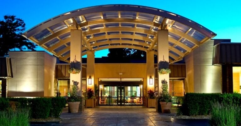 The image shows the entrance of a modern building with a large, curved glass canopy, illuminated by warm lights. Potted plants and neatly trimmed hedges line the pathway leading to the glass doors.