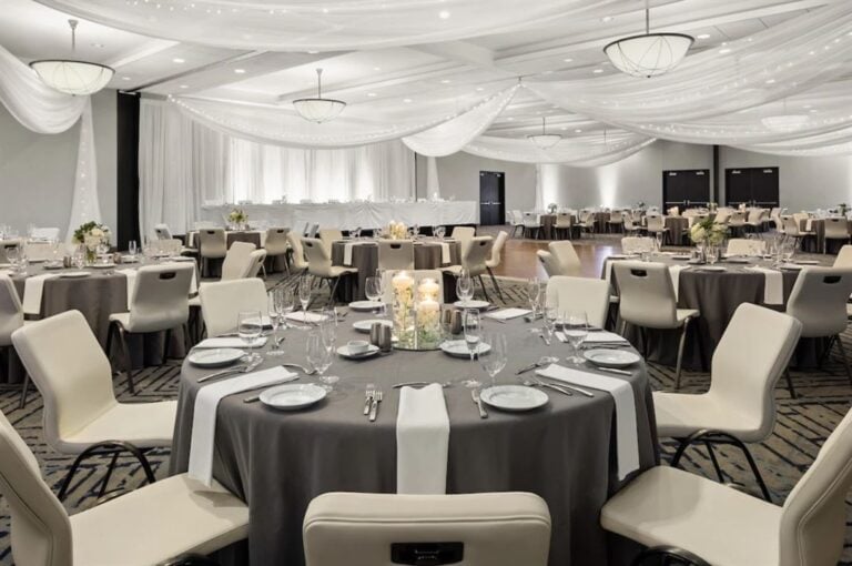A spacious banquet hall set for an event, with round tables covered in gray tablecloths and white runners, elegant place settings, cream chairs, floral centerpieces, and soft white drapes and lights hanging from the ceiling.