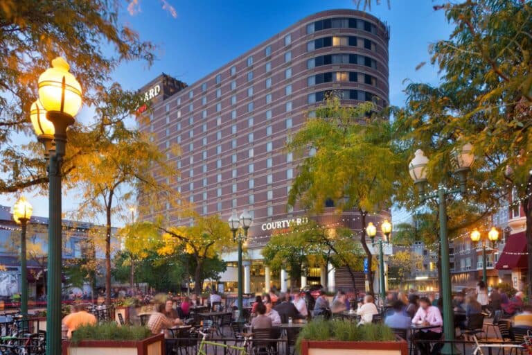 People dine at outdoor tables surrounded by trees and lamplight in a lively plaza, with a tall, modern Courtyard hotel building in the background at dusk.