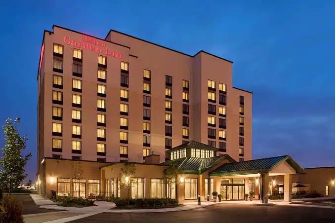A multi-story Hilton Garden Inn hotel building at dusk, with illuminated windows and a covered entrance, set against a clear blue sky.