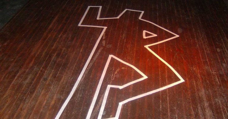 A white tape outline of a human figure is on a dark wooden floor, commonly used to indicate a crime scene. The outline suggests a person with one arm raised and legs slightly bent.
