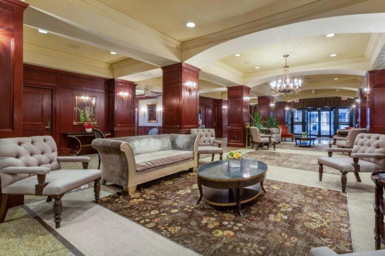Elegant hotel lobby with plush sofas, armchairs, glass coffee tables, chandeliers, polished wood paneling, decorative rugs, and plants. Large windows and glass doors are visible at the far end.