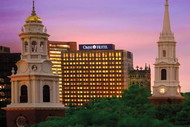 Two historic church towers with clock faces stand among green trees, with the brightly lit Omni Hotel building and a colorful sunset sky in the background.