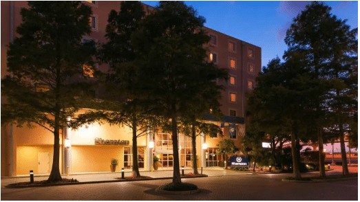 Exterior view of a Sheraton hotel at dusk, with warm lights glowing from windows and entrance, surrounded by tall trees and a clear sky transitioning from blue to pink.