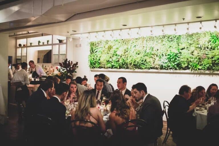 Guests in formal attire sit at tables, dining and conversing in a warmly lit restaurant. A lush green vertical garden decorates the wall, and staff are visible working near a bar area in the background.