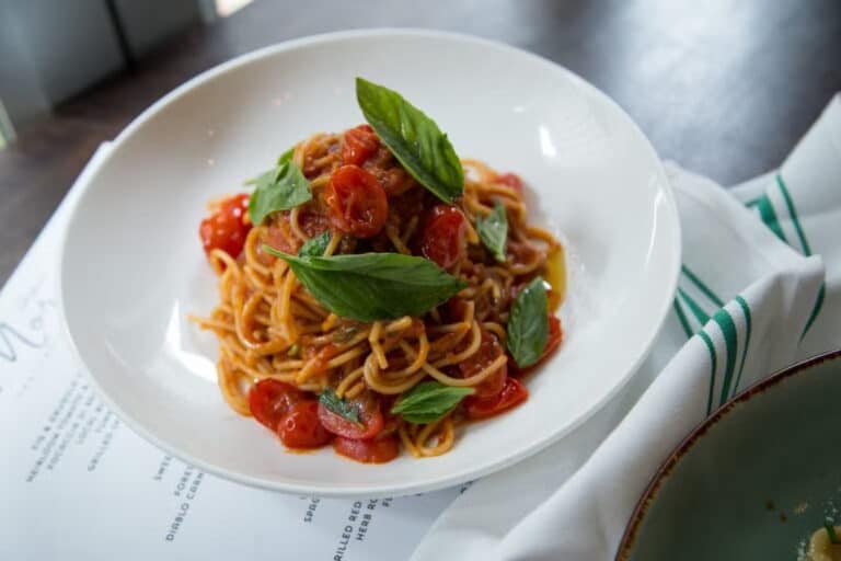 A plate of spaghetti topped with tomato sauce, sliced cherry tomatoes, and fresh basil leaves, served on a white dish next to a green-striped napkin.