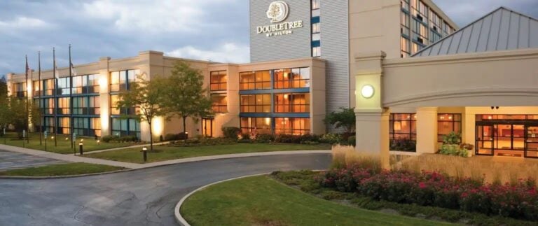 Exterior view of a DoubleTree by Hilton hotel, featuring a modern building with large windows, landscaped gardens, flags, and a driveway leading to the entrance, taken in the early evening.