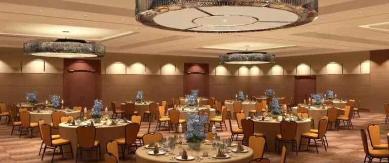 A spacious banquet hall with round tables set for an event, featuring yellow chairs, elegant table settings, blue floral centerpieces, and large modern chandeliers hanging from the ceiling.