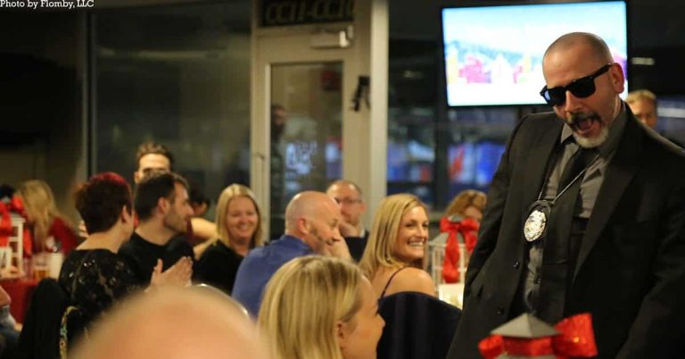 A man in a suit and sunglasses stands confidently in front of a seated audience at an indoor event. The audience is smiling and clapping. A large screen and festive decorations are visible in the background.
