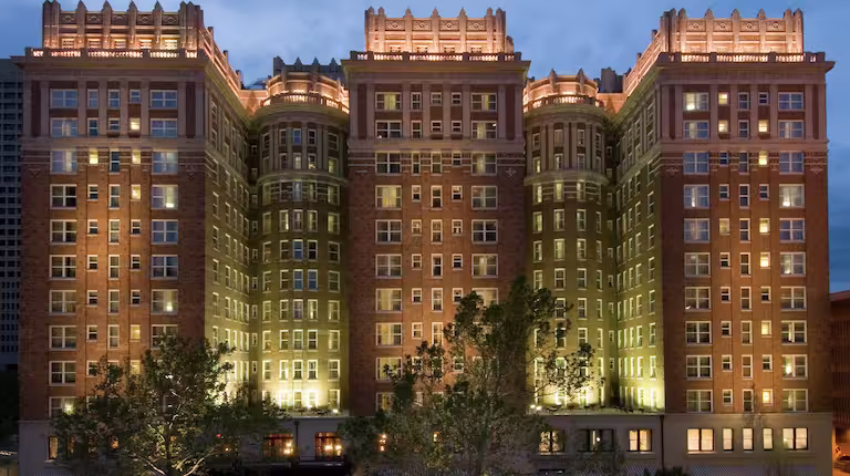 A large, historic brick hotel building with tall, illuminated windows and ornate architectural details is lit up at dusk, with trees in the foreground.