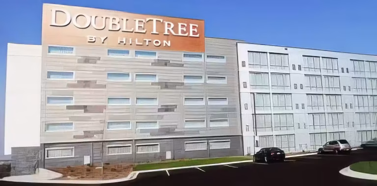A DoubleTree by Hilton hotel building with a large sign on the facade, multiple windows, and a parking lot in front, under a clear blue sky.