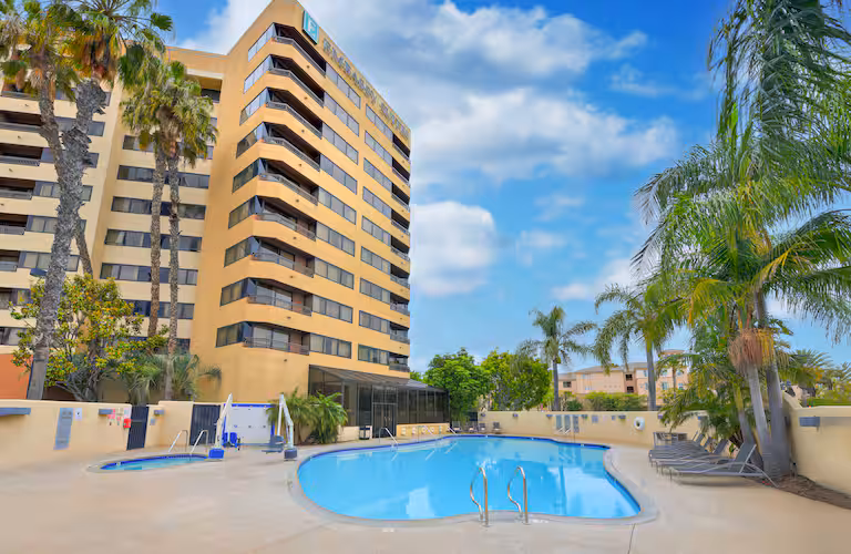 A modern hotel building with several floors stands beside a clear outdoor swimming pool, surrounded by palm trees, lounge chairs, and a bright blue sky with scattered clouds.