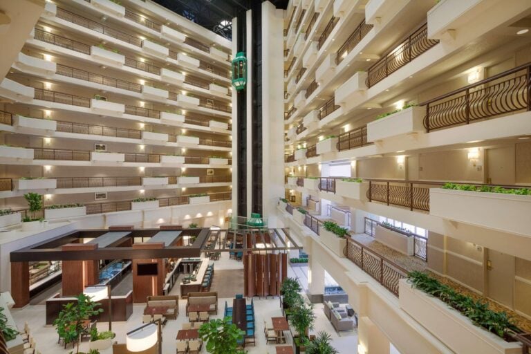Spacious hotel atrium with multiple floors of balconies, a glass elevator in the center, indoor plants, and modern seating areas on the ground floor under natural light.