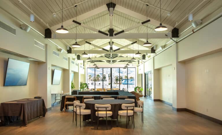 Spacious modern event room with high vaulted ceiling, round tables with chairs, large windows showing a mountain view, and natural light filling the space.