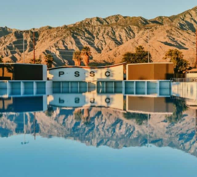 A clear swimming pool reflects palm trees, mountains, and buildings with the letters P S S C on them under a blue sky, creating a mirror image effect.