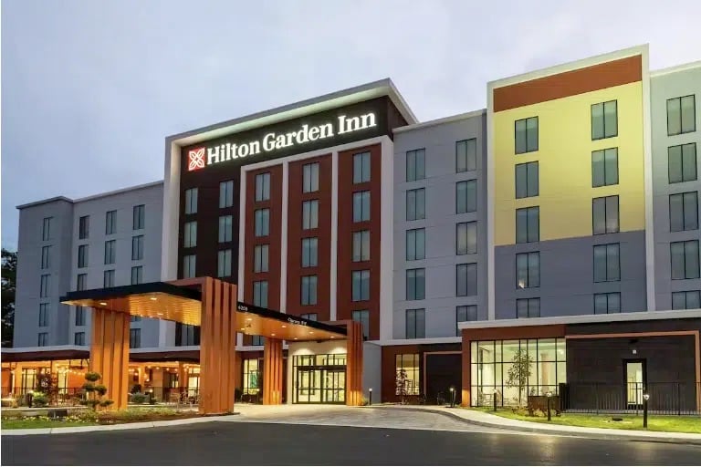 Modern exterior view of a Hilton Garden Inn hotel at dusk, with illuminated signage and large windows. The building features a covered entrance, landscaped grounds, and multiple stories.