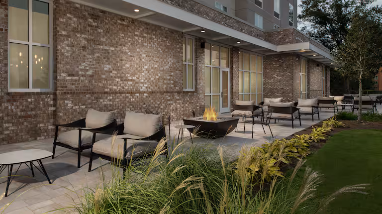 Outdoor patio with cushioned chairs, small tables, and a modern fire pit. The area is paved and bordered by plants, with a brick building and large windows in the background.