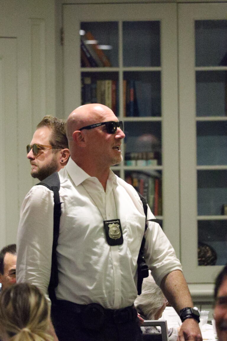 A bald man wearing sunglasses and a white dress shirt with a visible police badge in a shoulder holster stands in a room with bookshelves.