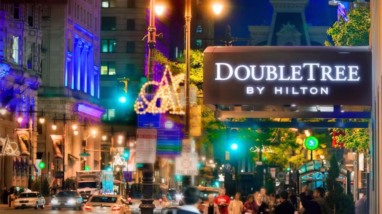 A lively city street at night with bright lights, people walking, and a prominent illuminated DoubleTree by Hilton hotel sign in the foreground. Buildings and festive decorations are visible in the background.