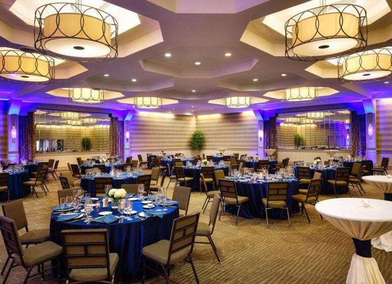 A spacious banquet hall set for an event, featuring round tables with navy blue tablecloths, elegant place settings, brown cushioned chairs, large decorative ceiling lights, and purple accent lighting.