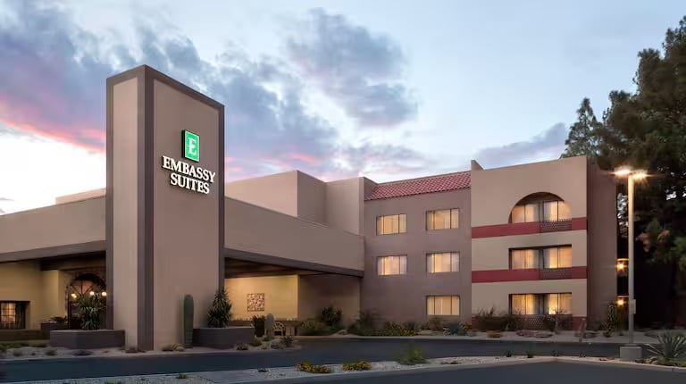 The image shows the exterior of an Embassy Suites hotel at dusk, with illuminated signage, beige and brown walls, large windows, and desert landscaping with cacti and small shrubs.