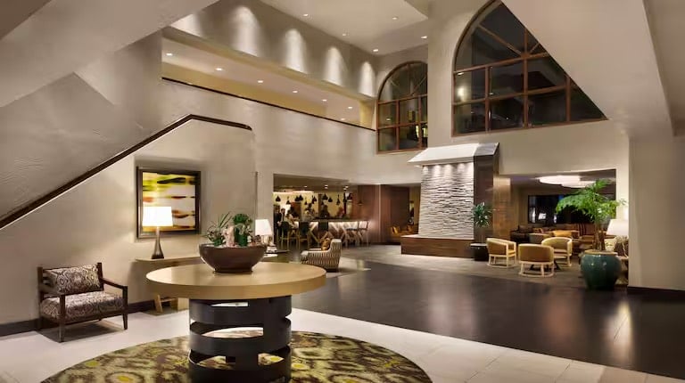 Modern hotel lobby with high ceilings, stylish furniture, large windows, and warm lighting. A round table with a decorative bowl sits on a patterned rug, and seating areas are arranged throughout the spacious, inviting room.
