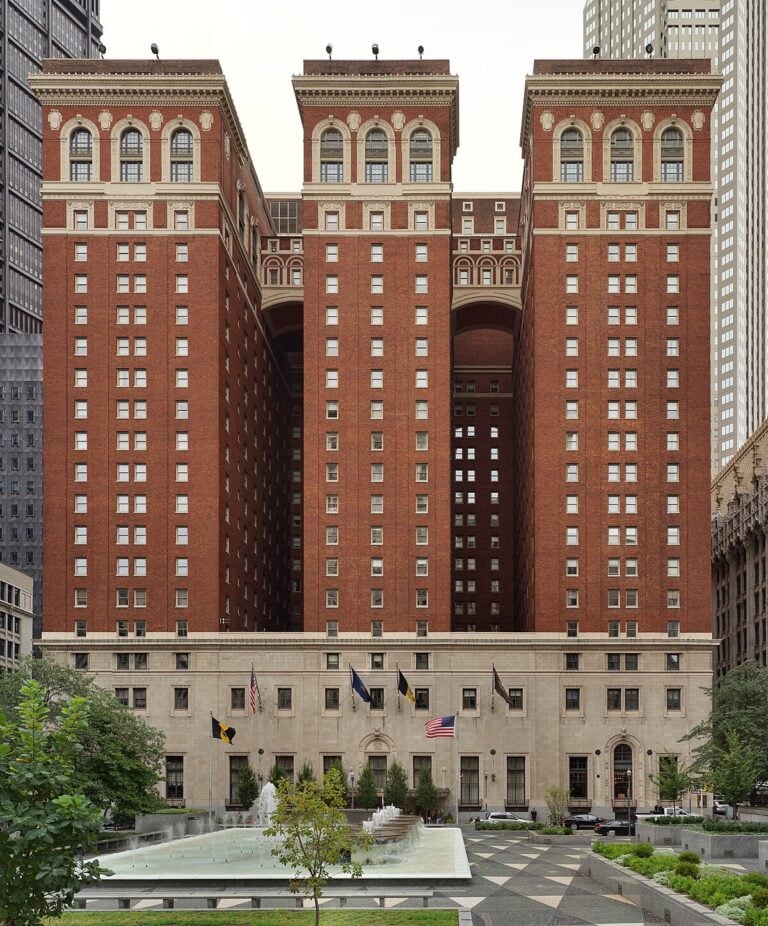 Symmetrical, historic red-brick hotel with three tall sections connected by arches at the top. The lower level features arched windows and multiple flags. A fountain and trees are in the foreground.