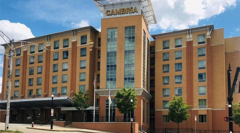 A six-story tan brick hotel with large windows and a modern glass central section. The sign at the top reads CAMBRIA. There are a few small trees, street lamps, and a blue sky with scattered clouds.